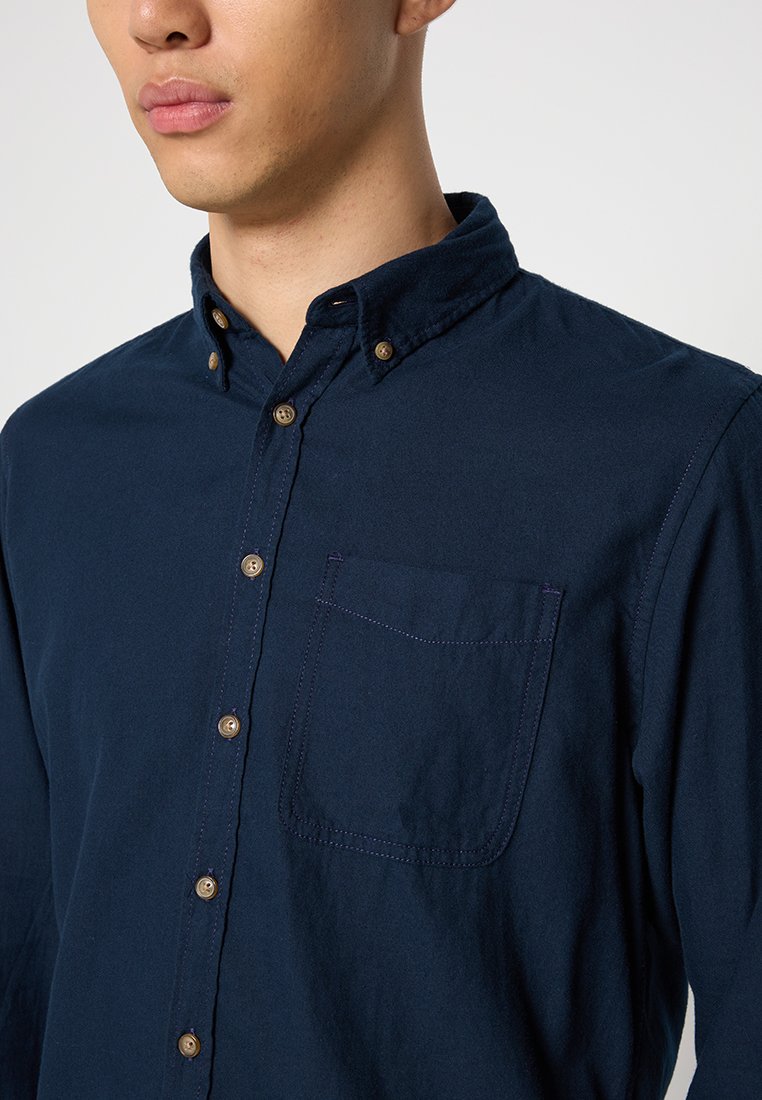 Camisa de botones azul marino hecha de algodón, con cuello, bolsillo frontal y botones marrones. Textura suave con detalles de costura sutiles.