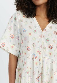 Bloemenpatroondress met een V-hals, korte mouwen en plooien. Gemaakt van lichte stof met kleurrijke ster- en bloemontwerpen op een witte achtergrond.