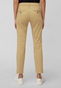 Pantalons beiges ajustés avec une coupe slim, dotés de deux poches au dos avec fermetures à bouton, fabriqués à partir d'un tissu lisse, associés à des baskets blanches.