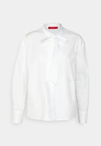 Blusa bianca con colletto a fiocco, maniche lunghe e chiusura frontale con bottoni. Presenta un dettaglio con logo ricamato sul lato, realizzata in tessuto morbido.
