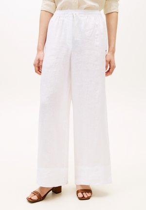 Donna che indossa pantaloni bianchi in lino a gamba larga con vita alta e coulisse, abbinati a sandali con tacco marroni e una camicia beige con maniche arrotolate.