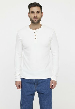 T-shirt henley à manches longues blanc en tissu texturé, avec trois boutons en bois au niveau du col, associé à un jean bleu.