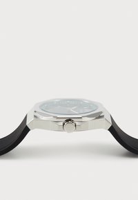 Octagonal klokke med et sølvfarget rustfritt stålkasse, svart gummirem og blågrå skive med timemarkører og krone-detalj.
