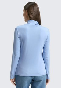 Vrouw met lang bruin haar, gekleed in een lichtblauwe coltrui en blauwe spijkerbroek, die met de rug naar de camera staat tegen een effen grijze achtergrond.