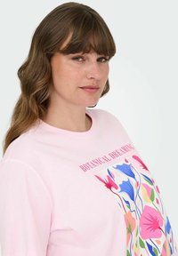Sieviete ar viļņainiem brūniem matiem valkā rozā krekls ar krāsainiem ziedu motīviem un tekstu "BOTANICAL DREAMING" uz baltā fona.