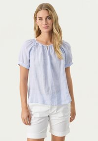 Himmelbluse mit kurzen Ärmeln und gerafftem Stoff am Ausschnitt sowie einer kleinen Öffnung vorne, kombiniert mit weißen Shorts.