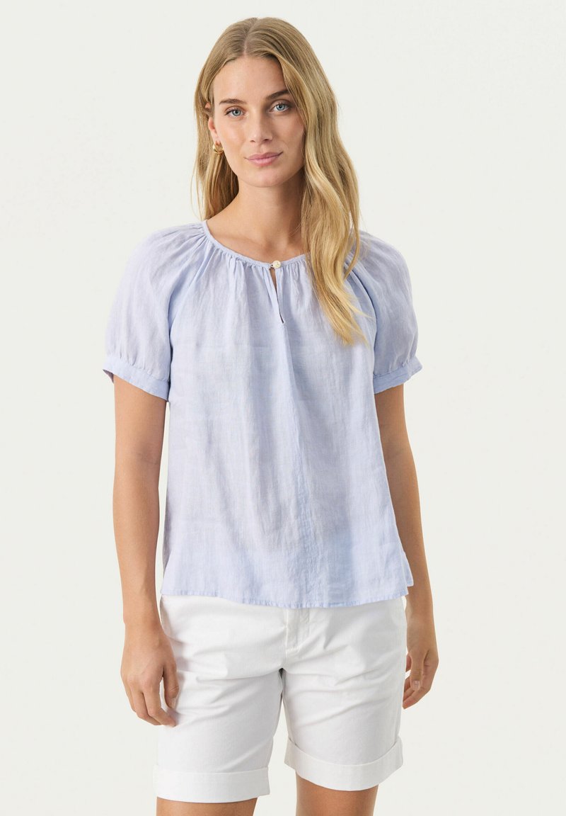 Light blue, short-sleeve blouse with gathered fabric around the neckline and a small opening at the front, paired with white shorts.