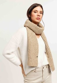 Sciarpa in maglia beige con un motivo a coste testurizzato, appoggiata attorno al collo di una persona che indossa un maglione crema. La sciarpa pende fino a metà torace.