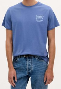 Tricou din bumbac albastru, cu mâneci scurte, având un grafic cu pește și text pe pieptul stâng. Purtați cu blugi decolorati.