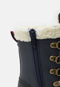 Botte imperméable bleu marine avec doublure en fausse fourrure, œillets en métal doré et détail de fermeture éclair sur le côté. Texture extérieure lisse et construction robuste.