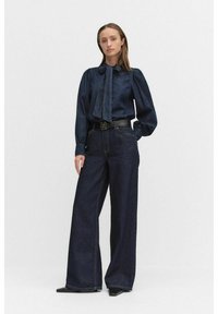 Mörk denimskjorta med puffärmar och knytning, kombinerad med vida mörka jeans. Svart bälte med dekorativ spänne.