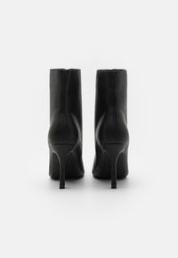 Paire de bottines en cuir noir à talons hauts et fins, vues de dos sur un fond blanc.