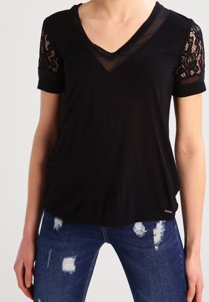 Sort T-shirt med V-udskæring og gennemsigtig meshpanel, med blondeærmer. Sammen med slidte blå jeans med synlige lapper.