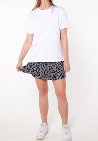 Bílý bavlněný tričko spárováno s černou sukní, která má bílé květinové vzory. Modelka nosí bílé tenisky a drží lem sukně.