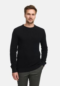 Svart, strikket genser med rund hals, lange ermer og ribbestrikkede mansjetter. Båret over teksturerte grå bukser. En enkel og tettsittende design.