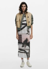 Giacca bomber leggera, beige e verde, con bottoni, abbinata a un lungo abito di seta stampato con motivi di palme e colori tenui.