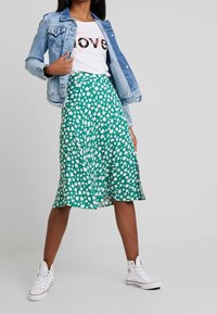 Groene bloemenrok met witte bladerenpatronen, gecombineerd met een eenvoudig wit t-shirt en een lichtblauwe spijkerjack. Witte sneakers maken de look compleet.