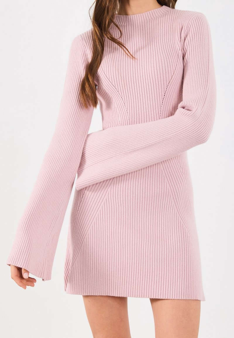Femme portant une robe en maille côtelée rose clair avec de longues manches cloche et une taille cintrée, montrée du cou jusqu'à mi-cuisse.