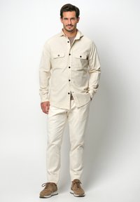 Beige overhemd met knopen en twee borstzakken, gecombineerd met crèmekleurige broeken. Schoenen zijn lichtbruin met witte accenten. Geverfde stof.