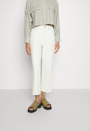 Personne portant un pantalon blanc large et court, une chemise courte à carreaux, et des sandales compensées tissées jaunes et noires.