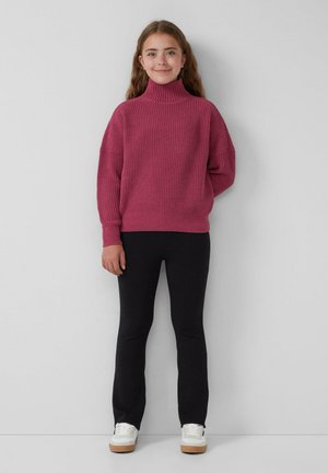 Rosa Strick-Turtleneck-Pullover mit weiten Ärmeln und geripptem Muster, kombiniert mit schwarzen enganliegenden Hosen und weißen Turnschuhen mit braunen Sohlen.