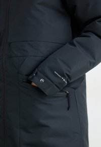 Hånd i den lynlåsede lomme på en sort Omni-Tech jakke med trykknap på manchetterne, der viser stoftekstur og branding på ærmet.