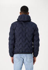 Giacca puffer blu navy con motivo a chevron, grande cappuccio e polsini elasticizzati, abbinata a jeans in denim chiaro.