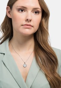 Srebrny naszyjnik z okrągłym wisiorem zdobionym misternym grawerunkiem kwiatowym, noszony na jasnobrązowej marynarce, z widocznymi średniej długości falowanymi włosami.