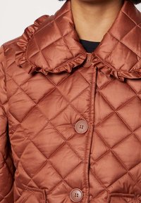 Pikowana kurtka o rdzawym kolorze z falbankowym kołnierzem, dwoma guzikami i bocznymi kieszeniami. Gładka, błyszcząca tekstura z diamentowymi wzorami.