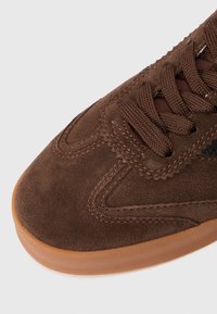 Smeđi nubuk tenisice s glatkom teksturom, koje imaju sukladne smeđe vezice, prošivene detalje i svjetlosmeđi gumeni potplat.