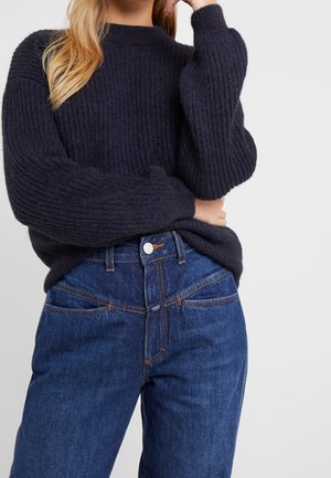 Femme portant un pull en grosse maille noire et un jean bleu taille haute, avec un bras plié et la main près du visage.