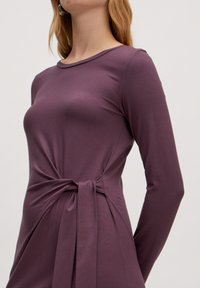 Robe à manches longues en mauve profond, réalisée en tissu doux, présentant un détail de nœud tordu à la taille pour plus de texture et de structure.
