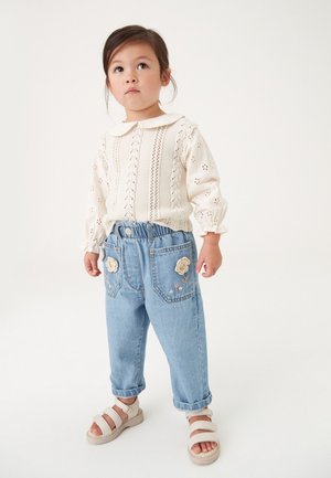 Weiße gestrickte Bluse mit Kragen, leichte blaue Jeanshose mit floralen Stickereien und beigen Sandalen mit mehreren Riemen.