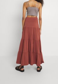 Jupe maxi couleur rouille avec un design à volants, fabriquée en tissu doux. Associée à un haut court gris et des sandales noires à lanières.