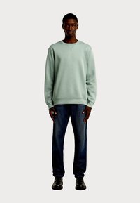 Jasnozielony sweter z okrągłym dekoltem i długimi rękawami, wykonany z teksturowanej tkaniny, z małym logo, zestawiony z ciemnoniebieskimi jeansami i czarnymi butami.