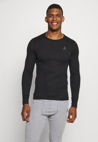 Schwarzes langärmliges Thermoshirt mit rundem Ausschnitt und taillierten Design, mit einem dezenten Logo auf der Brust und strukturiertem Stoff. Graue Hose darunter.