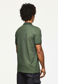 Mężczyzna z kręconymi włosami, ubrany w zieloną koszulkę polo i czarne spodenki cargo, stojący plecami do obiektywu na gładkim białym tle.