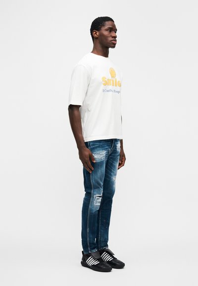Λευκό γραφικό μπλουζάκι με κίτρινο χαμογελαστό πρόσωπο και κείμενο, συνδυασμένο με φθαρμένο μπλε τζιν τζιν και μαύρα αθλητικά παπούτσια με λευκές ρίγες.