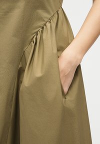 Χέρι τοποθετημένο στην πλαϊνή τσέπη ενός φορέματος από ελαφρώς μαζεμένο ύφασμα ελιάς, εμφανίζοντας υφή και λεπτομέρειες ραφής του φορέματος.