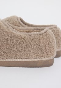 Een paar beige, zachte instapslippers met een gestructureerde stof en bruine rubberen zolen op een witte achtergrond.