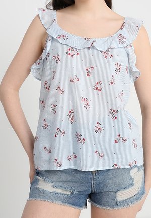 Kvinde iført en blå- og hvide stribet ærmeløs top med pink blomsterprint og flæsekant, kombineret med slidte denimshorts.