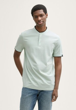 Světlé zelené polo tričko s henley límcem, třemi knoflíky a černým akcentem na límci; nošeno s modrými džíny. Hladká textura.