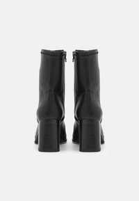 Une paire de bottines noires à talons carrés et fermetures éclaires à l'arrière, vue de dos sur un fond blanc.