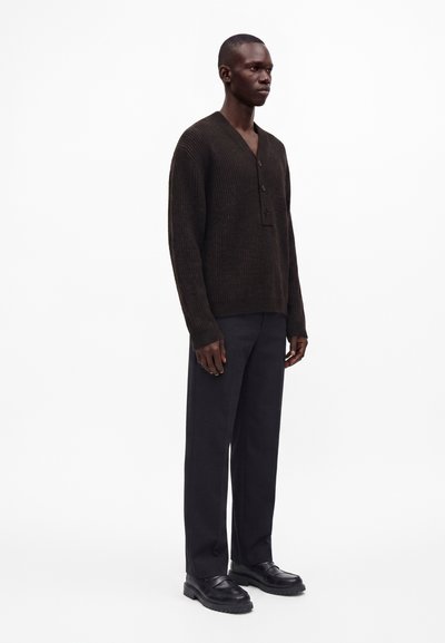 Homme debout portant un pull côtelé marron foncé boutonné, un pantalon noir et des chaussures en cuir noir sur fond blanc.