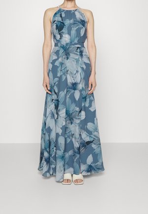 Kobieta ubrana w niebieską suknię bez rękawów sięgającą podłogi z dużymi białymi i niebieskimi kwiatowymi wzorami oraz białe buty z odkrytymi palcami.