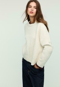 Cream-colored, textured sweater with a rounded neckline and ribbed hem, paired with dark denim jeans. The design is relaxed fit.