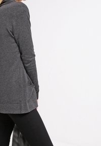 Cárdigan gris con capucha, mostrado desde la parte de atrás. Presenta bolsillos laterales y una textura suave. El dobladillo es ligeramente más largo en la parte posterior.