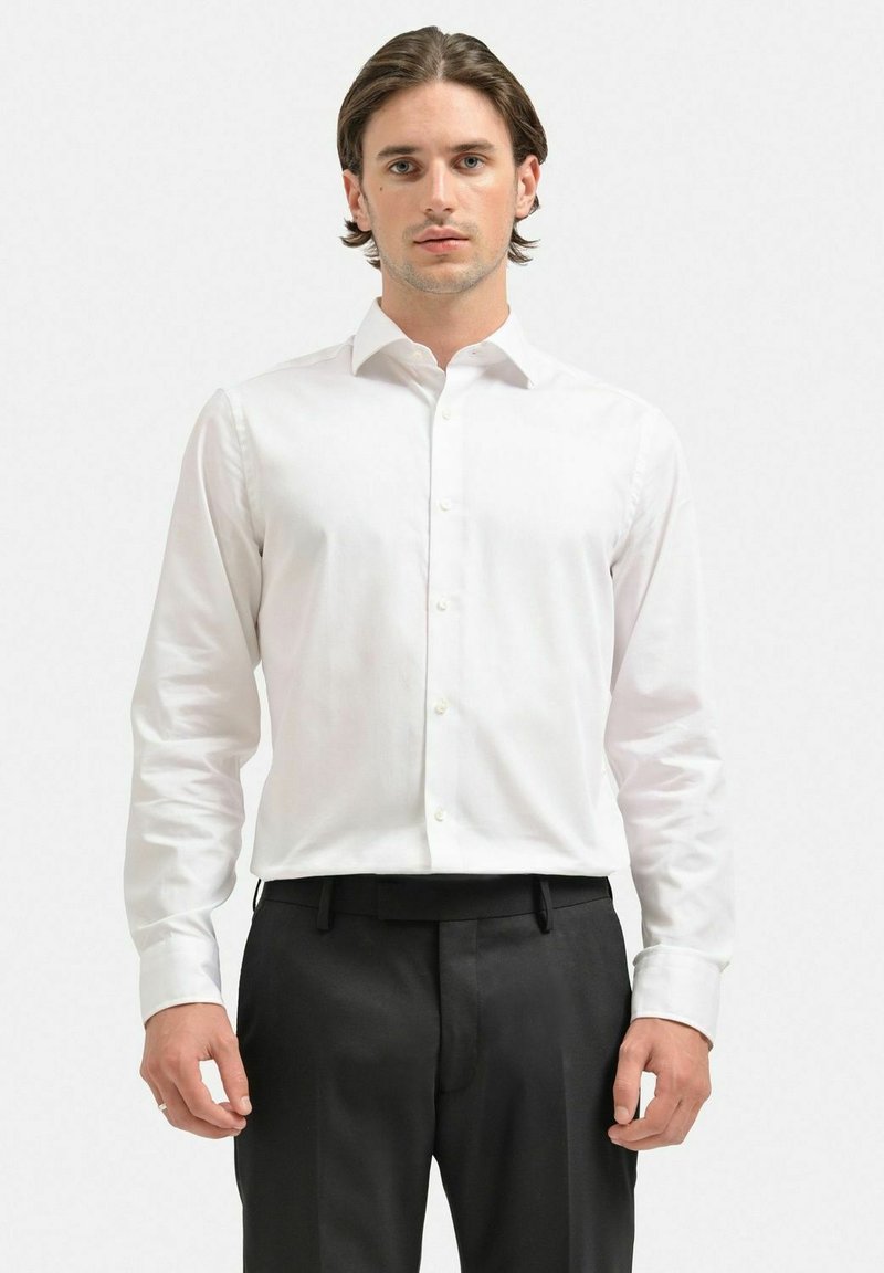 Vit långärmad skjorta med knappar, slim fit, mjukt tyg, klassisk krage, framplacket med knappar, bärs med svarta byxor.