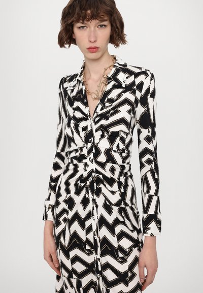 Rochie cu model alb și negru, cu guler, mâneci lungi și închidere cu nasturi. Prezintă un design în zigzag și o talie strânsă pentru structură.