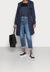 Persona che indossa un trench blu navy, un top blu scuro, jeans blu risvoltati, sneakers navy, mentre tiene una borsa a mano in pelle nera.